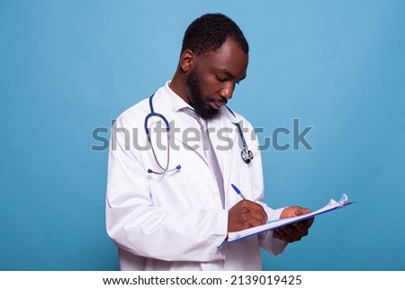 Portrait of health care medic in white lab coat holding clipboard writing on patient chart in clinical consult. Professional doctor with stethoscope taking notes looking at medical history.