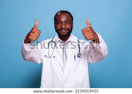 Portrait of cheerful african american doctor giving thumbs up in medical uniform with stethoscope looking at camera. Optimsitic medic showing hand gesture for positive attitude.