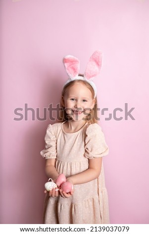 Happy Easter. A cute girl with rabbit ears holds colored eggs in her hands and smiles cheerfully. pink background. Space for text. High quality photo