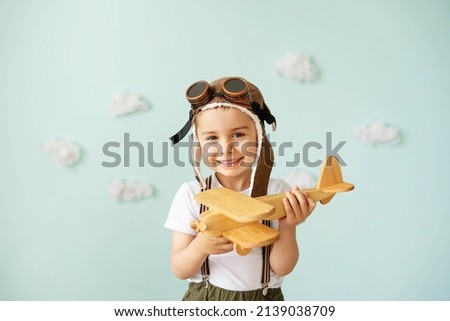 A boy in an aviator helmet and suspenders stands on a blue background with clouds and plays with a wooden plane. Royalty-Free Stock Photo #2139038709