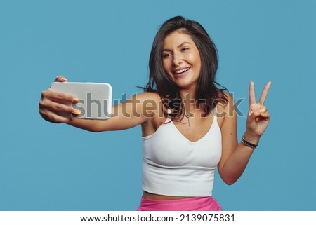 Stylish cheerful young woman showing peace gesture while having video-call, holding smart phone, wearing white top, isolated on blue background