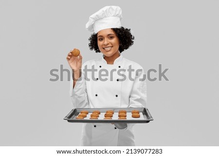 cooking, culinary and bakery concept - happy smiling female chef or baker in white toque and jacket holding baking tray with oatmeal cookies over grey background Royalty-Free Stock Photo #2139077283