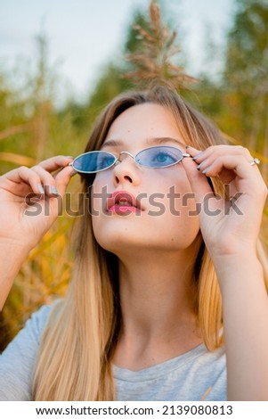 Beautiful young girl wearing blank gray t-shirt and black jeans in blue glasses posing against high green and yellow grass in early warm autumn. Outdoor portrait of beautiful female model. Vertical.