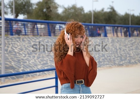 Young and beautiful red-haired woman listening to music with white headphones in a park where there is a big lake. The woman is happy. Concept expressions and happiness, music and dance.