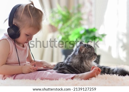 A little girl of two years old is sitting in black headphones and watching cartoons on her phone next to a gray cat.