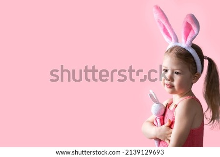 A little blonde girl in a pink romper with the Easter bunny ears on her head hugging a toy bunny. Close-up cute little girl with the Easter bunny ears on a pink background