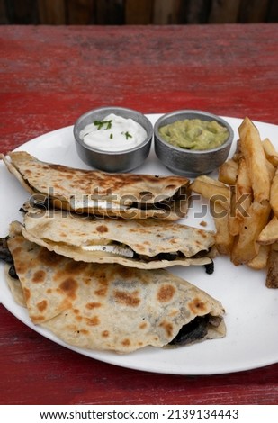 Mexican cuisine. Closeup view of quesadillas filled with cheese, french fries and dipping sauces, in a white dish on the restaurant red wooden table.