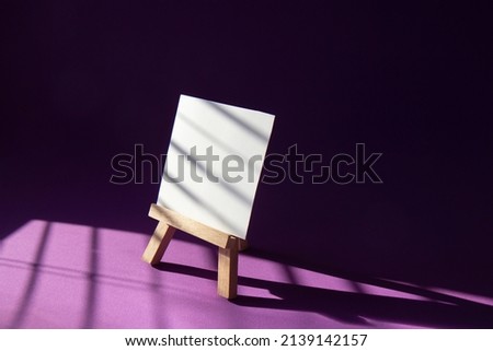 Wooden easel with a blank white sheet of paper on a bright purple background. Art concept