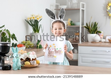 Cute little girl with bunny ears and Easter greeting card in kitchen at home