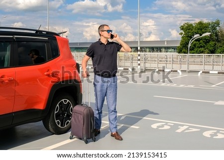 Young man standing with luggage near his car and talking on the phone outdoors. Travel concept.