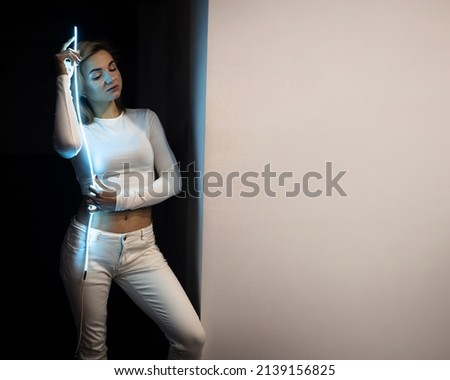 The portrait of beautiful woman in white clothes, top, shiny shimmer makeup and stars with blue led stripes, neon light on black background. Futuristic style girl, sadly emotions. Future, amazing.