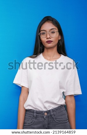 Closeup of Serious Young Indian Girl or Woman in white t shirt  and spects over a blue background. Attractive young teenager student girl from India  looking Straight at camera, posing for photo.