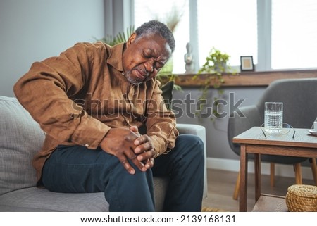 An African mature man suffers from knee pain while sitting on a couch. A mature man massages his aching knee. A man who suffers from knee pain at home, up close. Puts both hands on an aching knee. Royalty-Free Stock Photo #2139168131