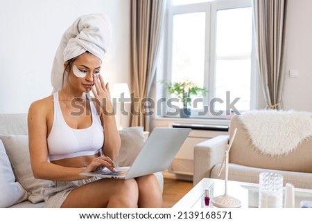 Side view in full length of a beautiful elegant lady in house clothes sitting on a bed with a laptop in her lap while using a mask for undereyes in the living room
