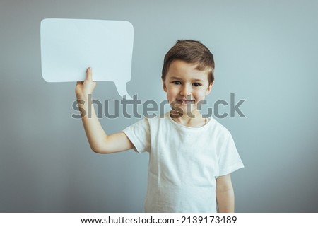 Smiling casual boy holding white paper message box with copy space for text above head looking at camera on gray background. Children speak loud concept