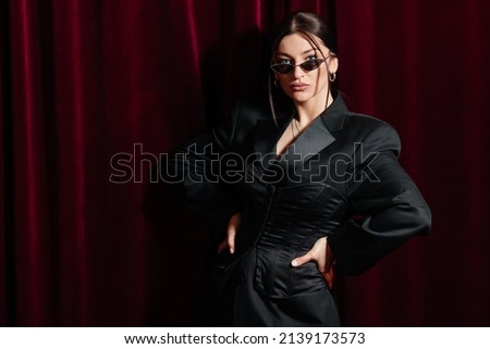 Photo of a model wearing all black posing in front of a red cinema curtain with nice sunglasses