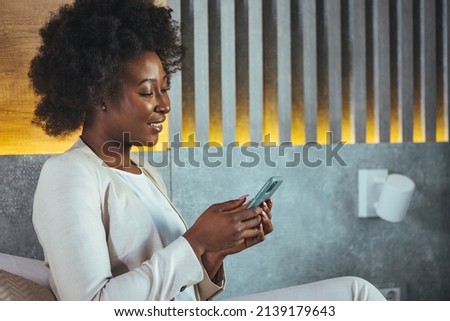 Shot of a young businesswoman using her cellphone in the hotel. Well Dressed young woman looking at her phone, checks and sends messages.Relaxing in bed in a hotel room after a trip

