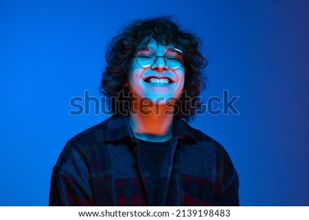 Looks happy, delighted. Portrait of young smiling man, student in black hoodie isolated on dark blue background in neon light. Concept of emotions, facial expression, youth, aspiration, sales. Royalty-Free Stock Photo #2139198483