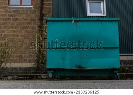 Blue green dumpster in front of tannish wall with turquoise building partially covering backdrop. Royalty-Free Stock Photo #2139236125