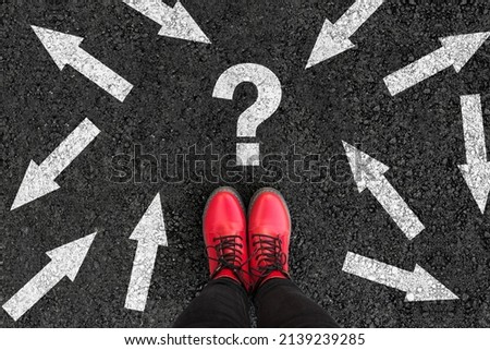 woman in shoes standing on asphalt next to multitude of arrows in different directions and question mark, confusion choice chaos concept  Royalty-Free Stock Photo #2139239285