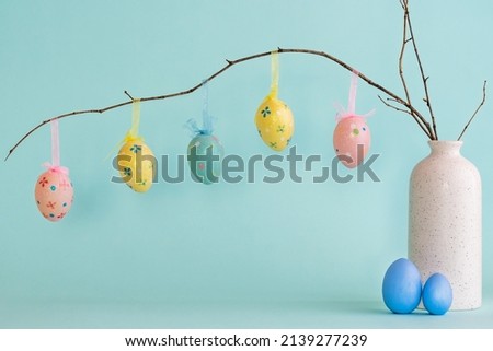 Decorative Easter eggs hanging on dry branch placed in white ceramic vase with two blue eggs near against blue background 