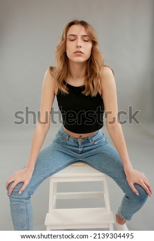 Beautiful girl model appearance in light jeans and black T-shirt on a gray studio background. Girl model sitting on a white chair. Beautiful blonde shows poses for a photo shoot