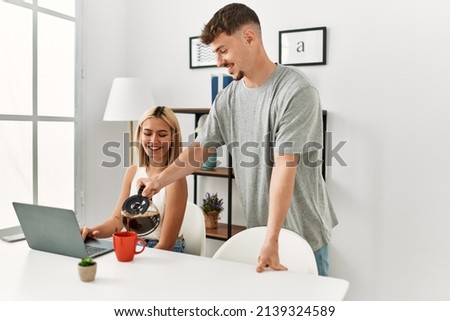 Woman working using laptop at home and her boyfriend pouring coffee.
