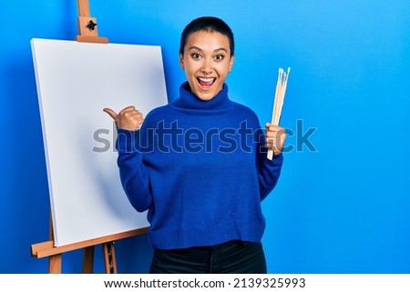 Beautiful hispanic woman with short hair holding brushes close to easel stand pointing thumb up to the side smiling happy with open mouth 