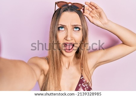 Young blonde girl wearing bikini taking a selfie angry and mad screaming frustrated and furious, shouting with anger looking up. 