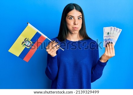 Young hispanic woman holding colombia flag and colombian pesos banknotes puffing cheeks with funny face. mouth inflated with air, catching air. 