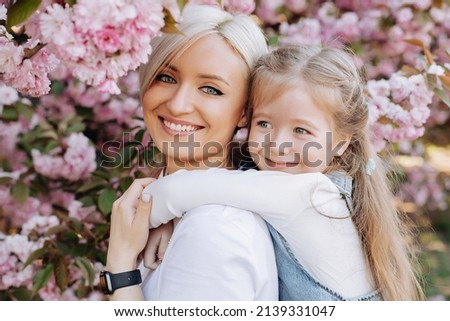 Close up of daughter hugging mom sitting on her back on cherry sakura background. Walk in the sakura trees in the city.