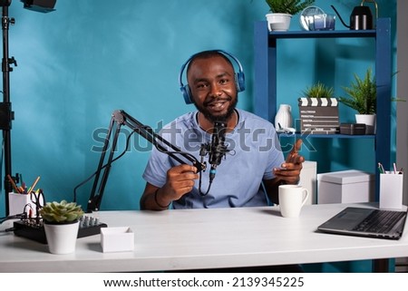 Smiling vlogger wearing headphones interacting with audience during online live show looking at camera sitting at desk with broadcasting setup. Influencer talking with fanbase in recording studio.