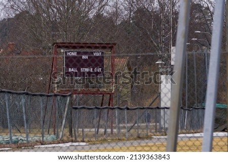 an old red rusty baseball score sign turned off in the suburbs from a far with fence in foreground