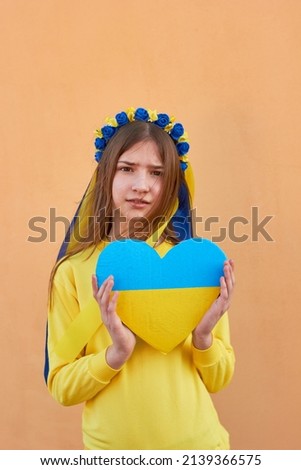 A little Ukrainian girl in a wreath on her head, holding a heart in the colors of the flag of Ukraine. Pray for Ukraine. Save Ukraine. The children are asking for peace. There is no war.