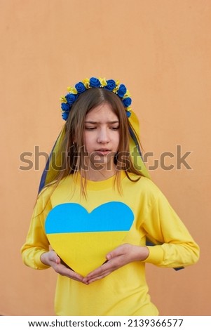 A little Ukrainian girl in a wreath on her head, holding a heart in the colors of the flag of Ukraine. Pray for Ukraine. Save Ukraine. The children are asking for peace. There is no war.