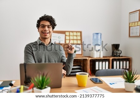 Young hispanic man wearing business style sitting on desk at office with a big smile on face, pointing with hand and finger to the side looking at the camera. 