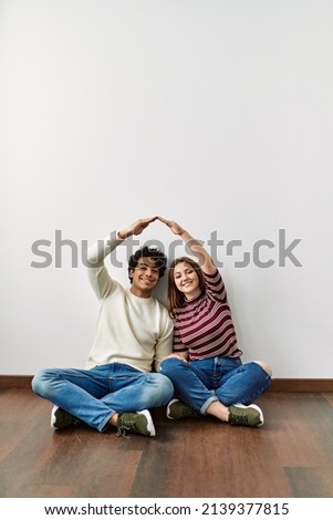 Young hispanic couple doing roof symbol with hands sitting on the floor at empty new home.