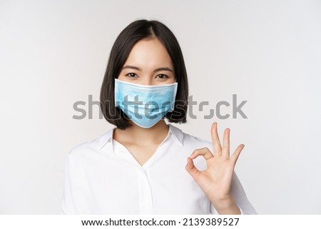 Covid and health concept. Portrait of asian woman wearing medical face mask and showing okay sign, standing over white background