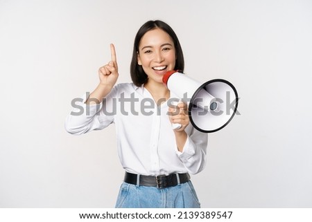 Smiling happy asian girl talking in megaphone and pointing up, announcing discount promo, showing advertisement on top, standing over white background Royalty-Free Stock Photo #2139389547