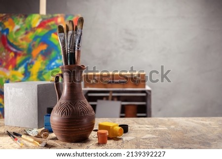 Artist supplies at wooden table. Paint brush in clay jug and art painter tool on desk background texture. Paintbrush for painting in artist workplace
