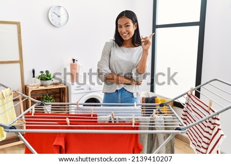 Young hispanic woman putting fresh laundry on clothesline with a big smile on face, pointing with hand and finger to the side looking at the camera. 