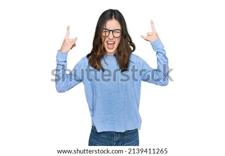 Young beautiful woman wearing casual clothes and glasses shouting with crazy expression doing rock symbol with hands up. music star. heavy music concept. 