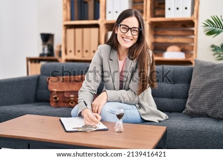 Young woman psychologist smiling confident writing on clipboard at psychology center Royalty-Free Stock Photo #2139416421
