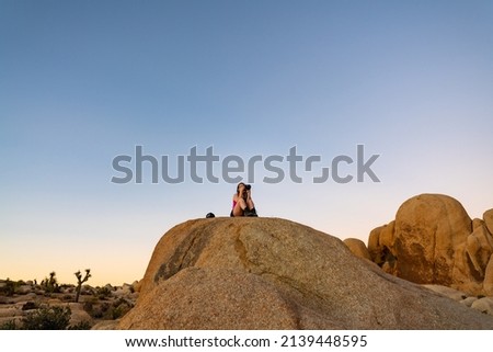 Woman tourist sitting atop a large boulder in Joshua Tree National Park at sunset with perfect background sky. 