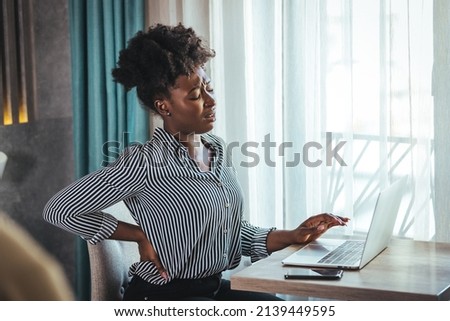 Tired african American millennial female worker sit at desk touch back suffer from lower spinal spasm, hurt unhealthy biracial woman stretch have strong backache, incorrect posture concept Royalty-Free Stock Photo #2139449595