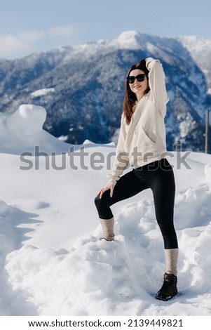  A young girl in a light sweater and dark leggings in glasses poses against the backdrop of mountains in winter and looks at the camera. High quality photo