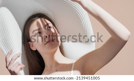 Young pretty European women with long brown hair in a big white hat covers face from the sun with her palm looking upward and holding the brim of her hat against beige background | Spf cream concept Royalty-Free Stock Photo #2139451005