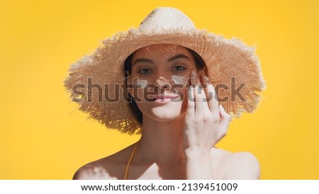 Young good-looking brunette Caucasian woman in a straw hat puts spf cream on her cheeks, enjoys the sun and smiles for the camera against yellow background | Spf cream concept Royalty-Free Stock Photo #2139451009