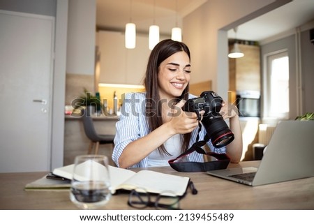 Happy young photographer holding a dslr camera in her home office. Female photographer smiling cheerfully while working at her desk. Creative female freelancer working on a new project. Royalty-Free Stock Photo #2139455489