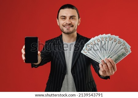 Young smiling successful rich latin man in black striped jacket grey shirt using mobile cell phone with blank screen area hold fan of cash money in dollar banknotes isolated on red background studio